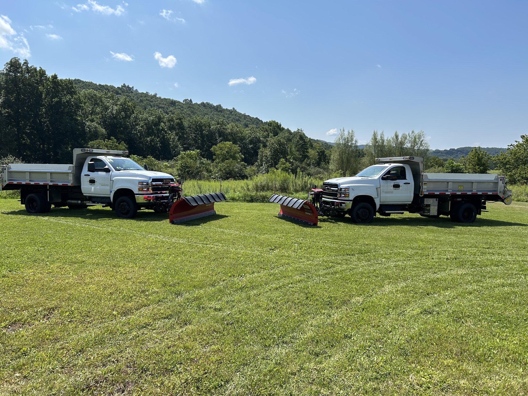 Two white Chevrolet 6500 municipal dump trucks in Snyder Township livery parked in a field, each fitted with a Galion stainless steel dump body and a red Western heavy-duty snowplow.